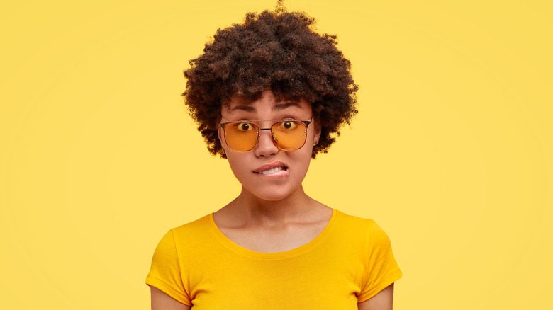 Женщина в очках и желтой рубашке кусает губу от смущения