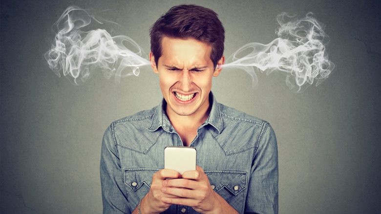 Сердитый мужчина с дымом, выходящим из ушей, смотрит на свой мобильный телефон