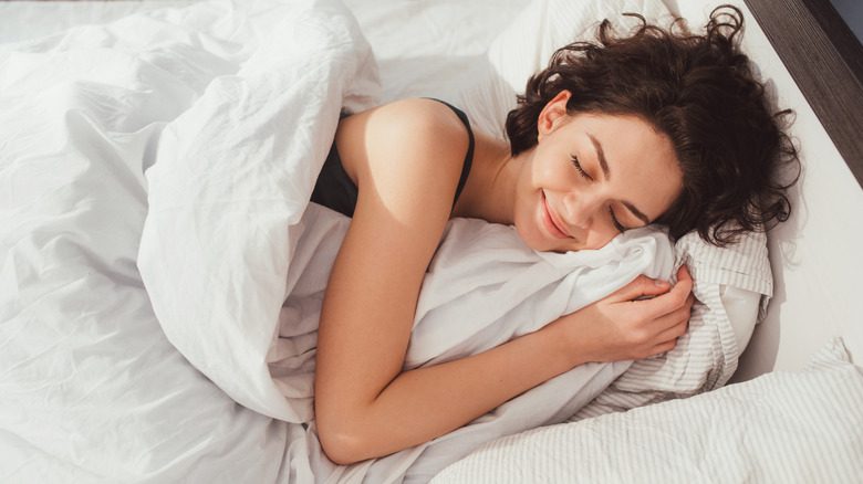 Улыбающаяся женщина лежит в постели с закрытыми глазами, укрытая белым одеялом.