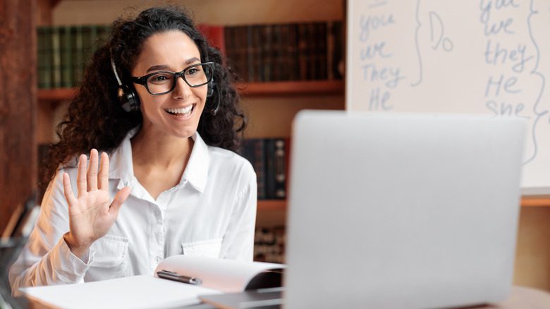 Сидящая за столом женщина в очках и гарнитуре машет рукой и улыбается экрану компьютера