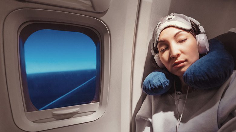 Женщина в наушниках, маске для глаз и подушке для шеи спит в своем кресле в самолете